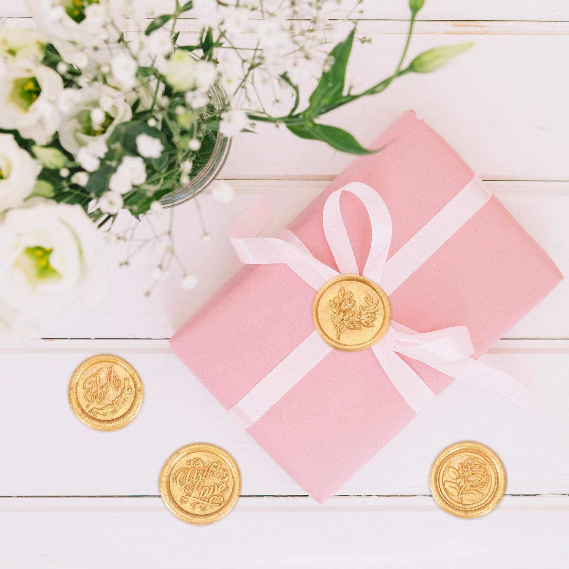 Pink gift box with a white ribbon and gold seals on a wooden surface with flowers in the background
