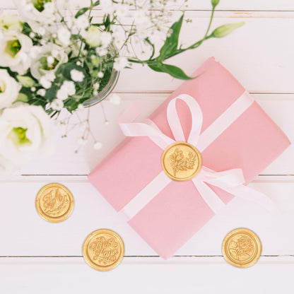 Pink gift box with a white ribbon and gold seals on a wooden surface with flowers in the background