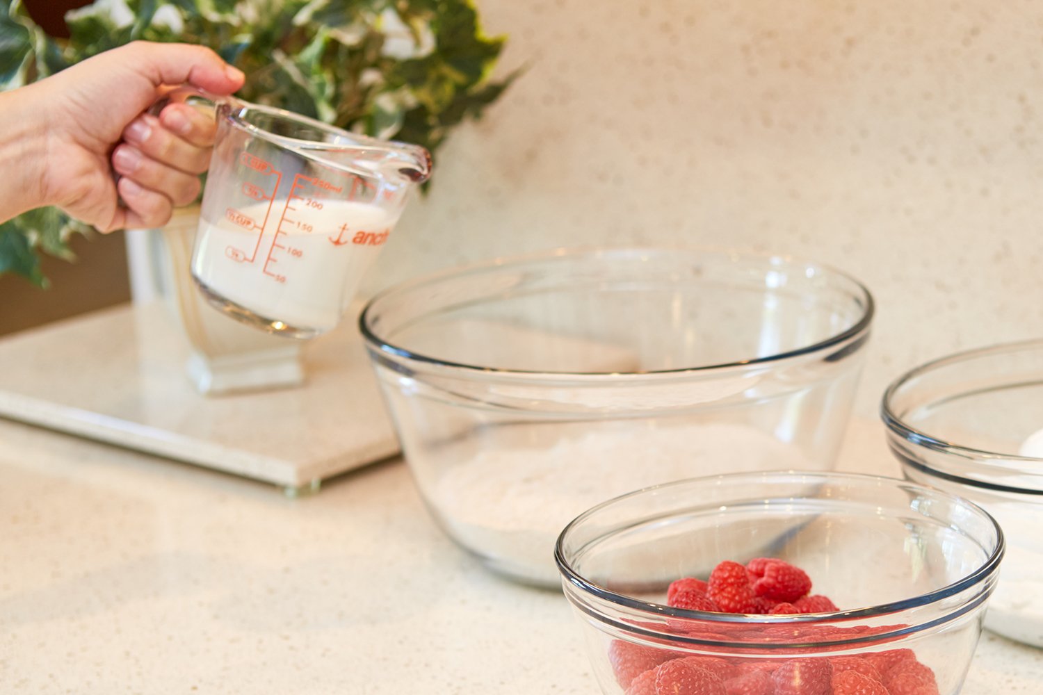 Hand holding a measuring cup with glass bowls and raspberries on a kitchen counter.