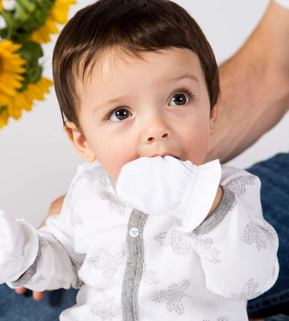 Baby wearing a white outfit with gray accents, sitting on a person's lap with sunflowers in the background.