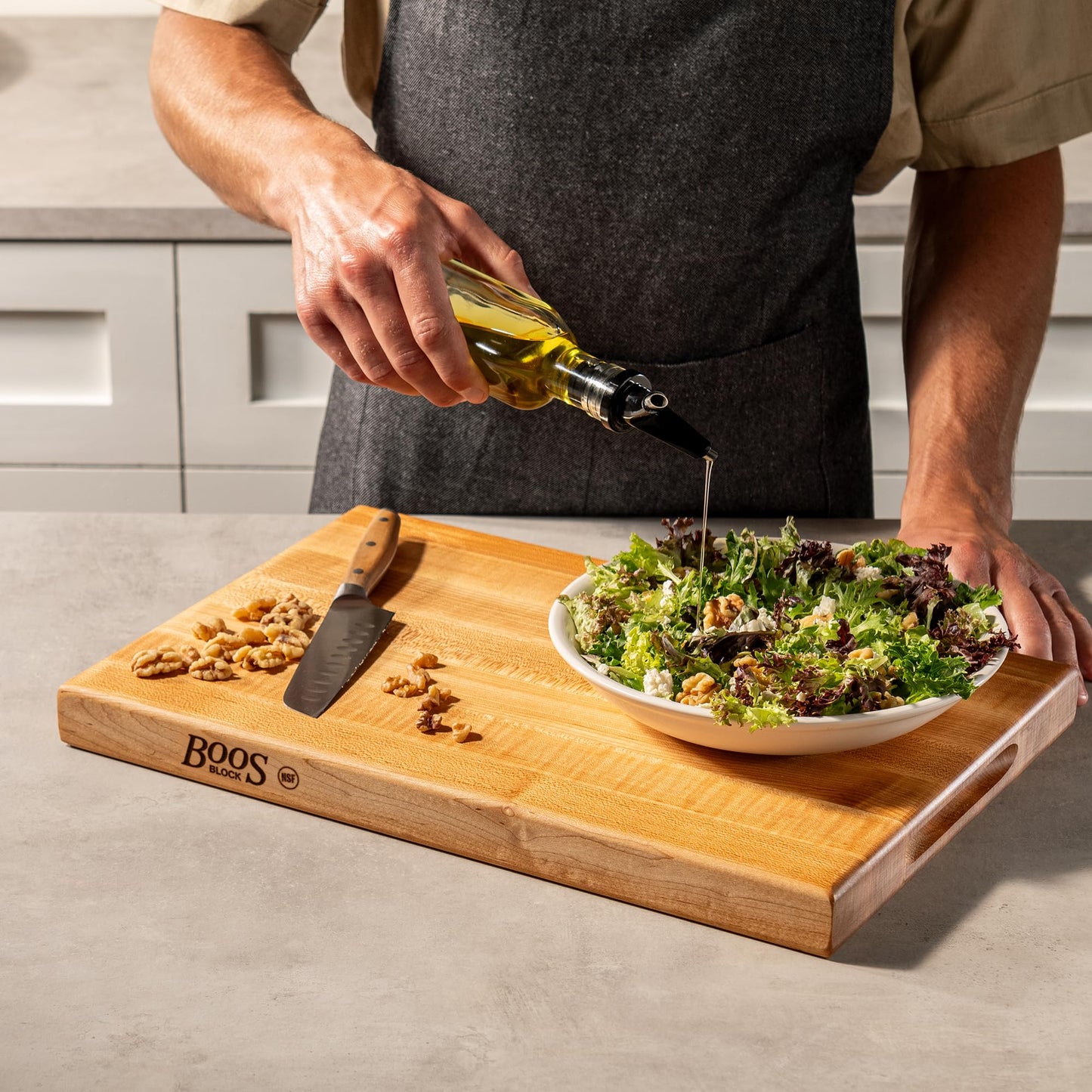 Person drizzling olive oil over a salad on a wooden cutting board with a knife.