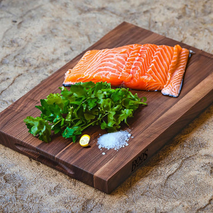 Raw salmon fillet with greens and salt on a wooden cutting board outdoors.
