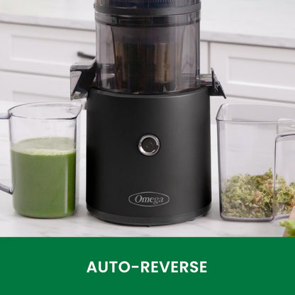 Black Omega juicer with a glass of green juice and a container of greens on a white countertop.