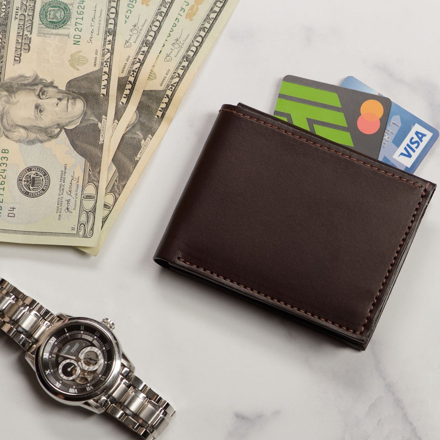 Brown leather wallet with cards, dollar bills, and a watch on a marble surface