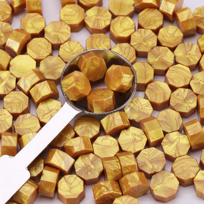 Gold-colored hexagonal sealing wax pieces with a metal scoop on a white background