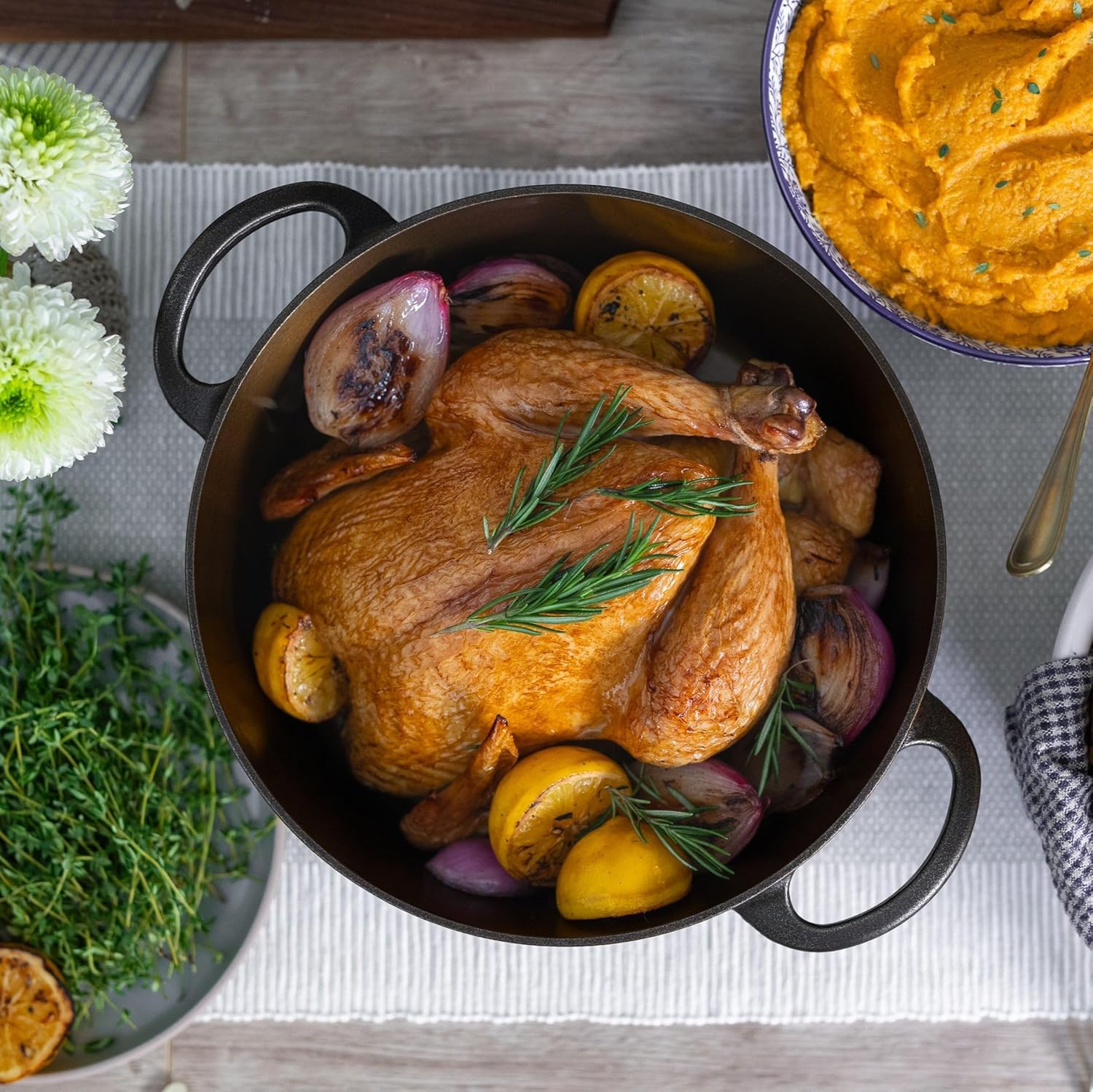 Roasted chicken with vegetables in a black pot on a table with flowers and mashed potatoes.