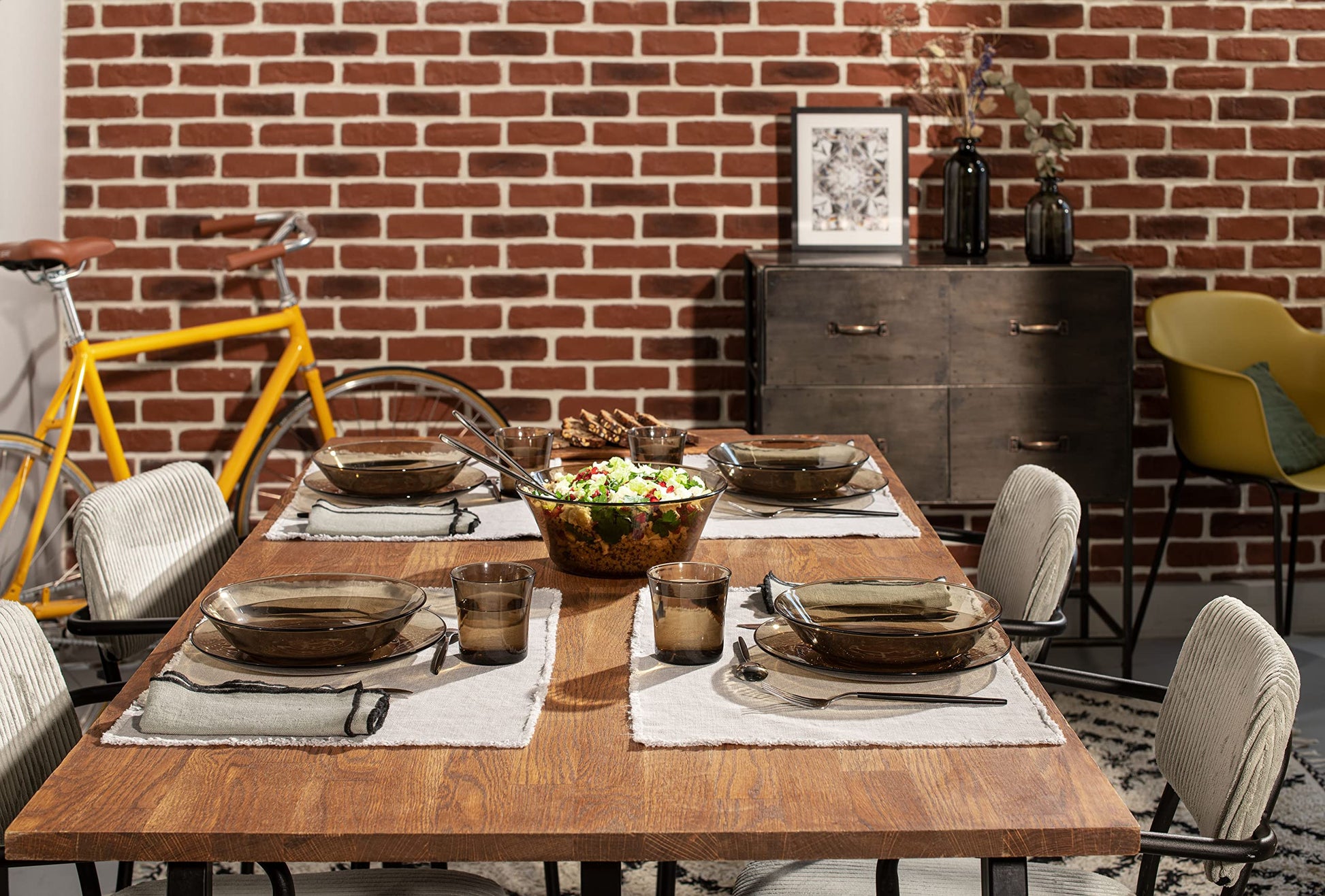 Dining table set with Duralex Lys Clear Glass Dinnerware Set, 18-Piece (Service for 6) against a brick wall with a bicycle and furniture in the background.