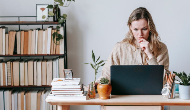 Woman sitting at a desk with a laptop, surrounded by books and plants.
